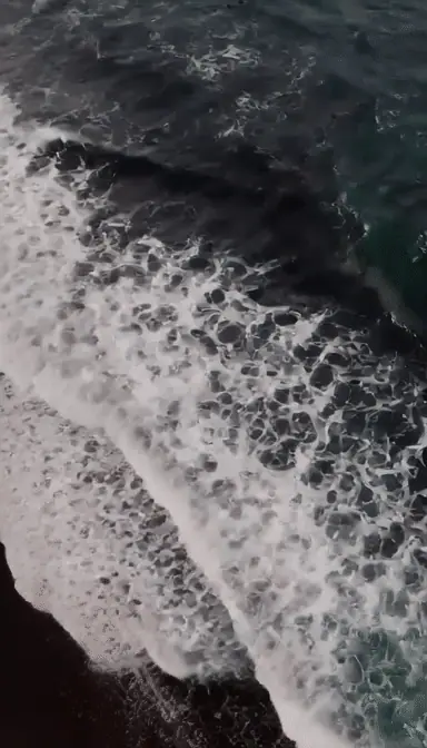 aerial view of turquoise ocean waves rolling onto a black sand beach, white foam